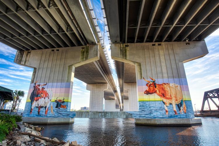 An overpass above a river there is a mural painted on the pillars supporting the structure in Jacksonville, Florida.